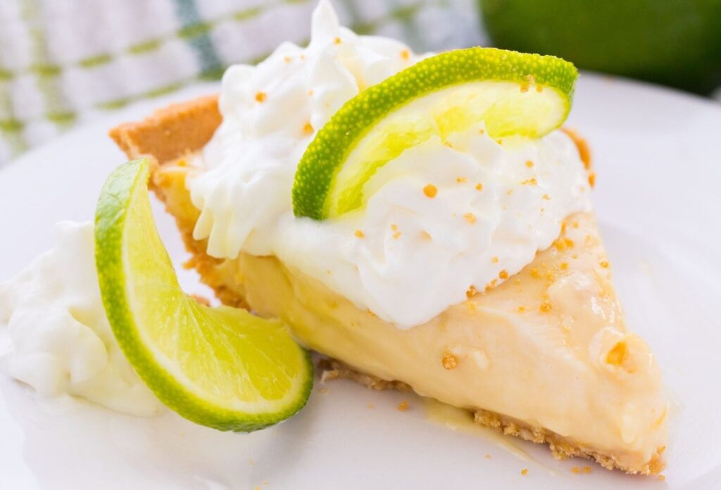 A slice of key lime pie topped with whipped cream and two slices of fresh lime, served on a white plate with an extra dollop of whipped cream beside it.
