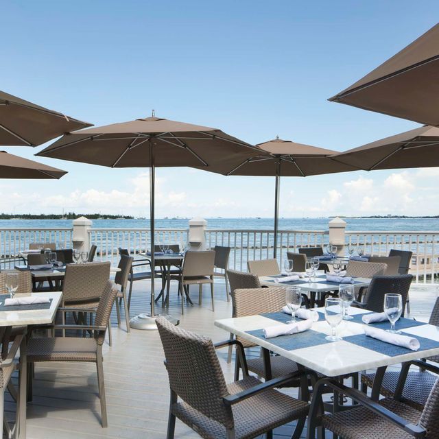 The ultimate marathon fl restaurants scene: from casual to fine dining 2 Outdoor restaurant patio with tables and chairs under large brown umbrellas, overlooking a calm ocean or bay on a sunny day. Blue sky and distant land are visible beyond a white railing. Tables are set for dining.