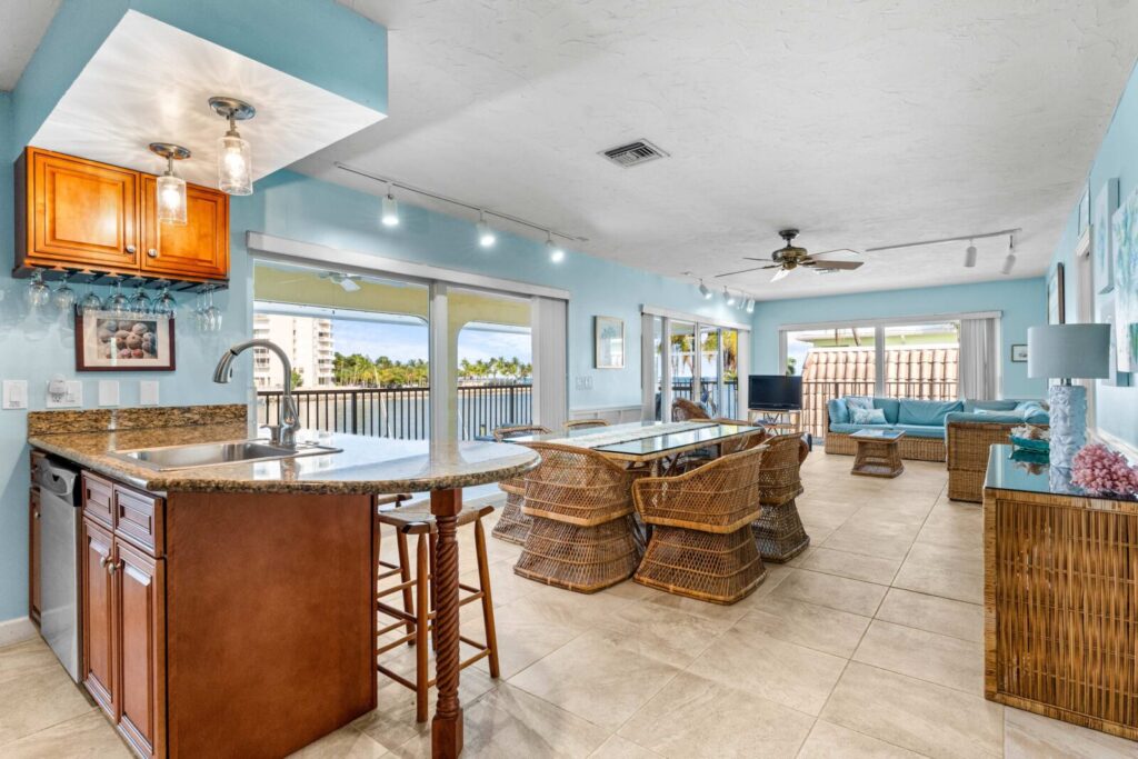 Open-concept kitchen and living area with wicker furniture, a granite island, light blue walls, and large windows offering a view of a balcony and palm trees outside. Natural light fills the airy, coastal-themed space.