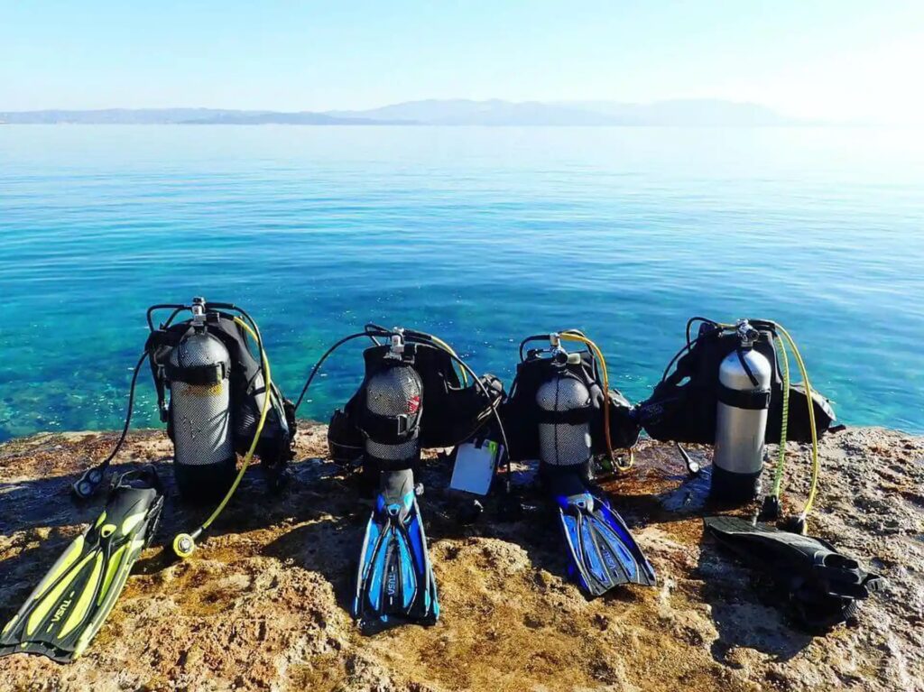 Four sets of scuba diving gear, including tanks, masks, and fins, are lined up on a rocky shore with clear blue water and distant mountains visible in the background under a clear sky.