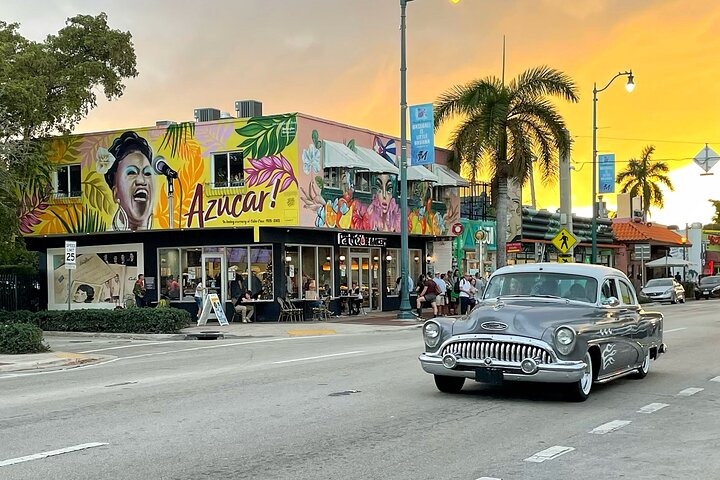 A classic gray car drives past a colorful mural featuring a singing woman and the word azucar! On a building at sunset, with palm trees and people dining outside on the sidewalk.