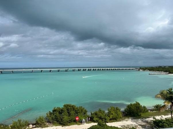 A wide view of turquoise ocean water under a cloudy sky, with a long bridge stretching across the water in the distance. Lush green trees and a small sandy shoreline are visible in the foreground.