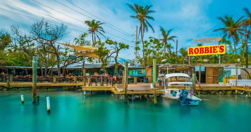 8 day trips from marathon fl you didn't know existed (2026) 11 Waterfront view of a tropical marina with a dock, small boats, palm trees, outdoor seating, and a large yellow and red sign that reads robbies under a blue sky.