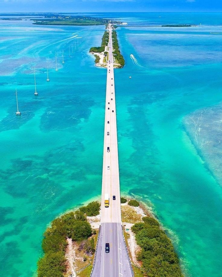 A long, narrow bridge stretches over clear turquoise water, connecting two pieces of land. Cars drive along the bridge, with sailboats anchored in the water and lush greenery at both ends. The sky is clear and blue.