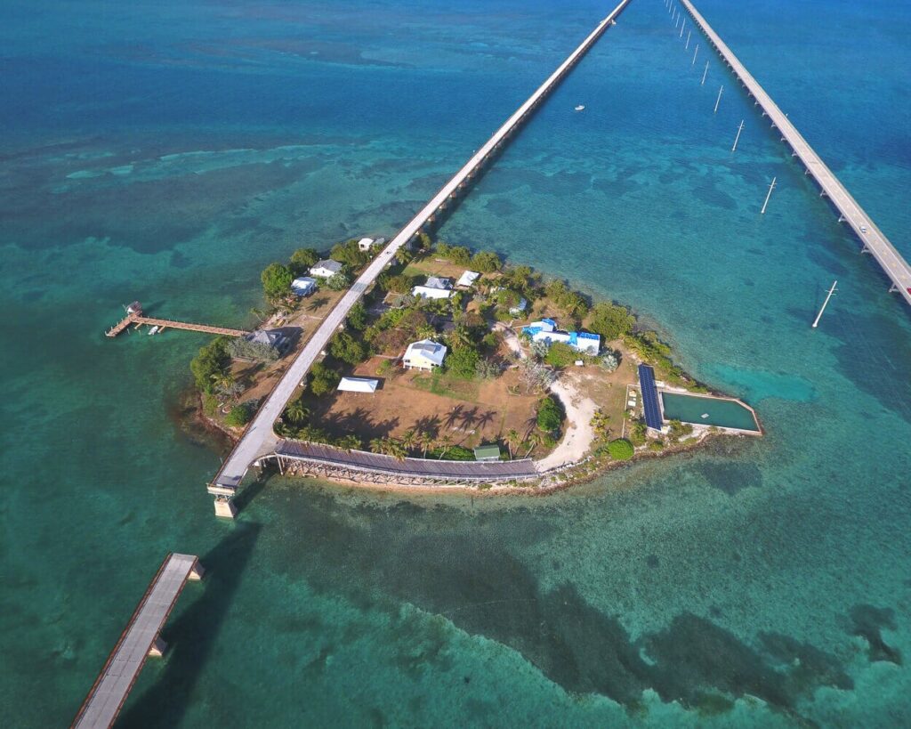 A small island with several buildings and trees is connected by two long bridges over clear turquoise water. The island is surrounded by shallow reefs, and a dock extends from its shore.