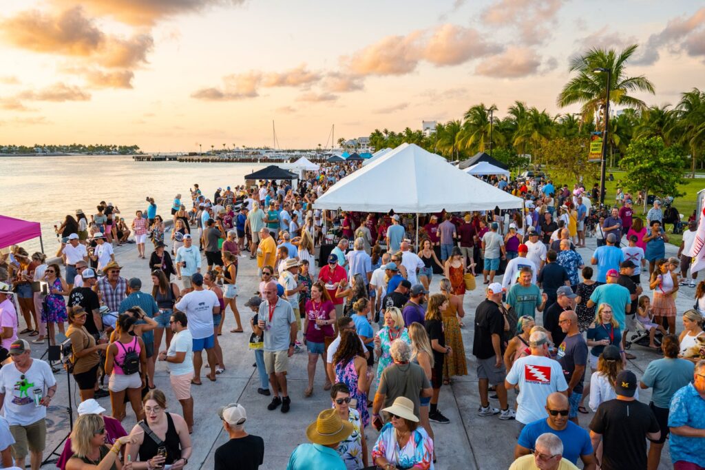 A large crowd gathers by the waterfront at sunset, with people socializing under white tents. Palm trees and boats are visible in the background, creating a lively, tropical atmosphere.