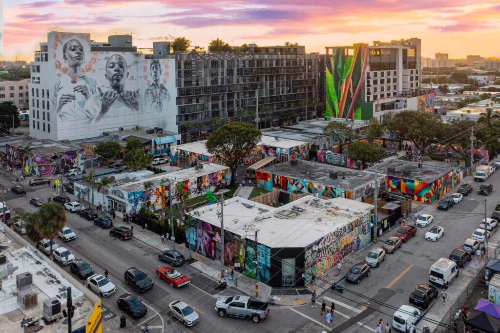A vibrant urban scene at sunset with colorful street art and murals covering buildings, cars parked along the roads, and people walking in the wynwood neighborhood of miami. The sky glows with shades of orange and purple.