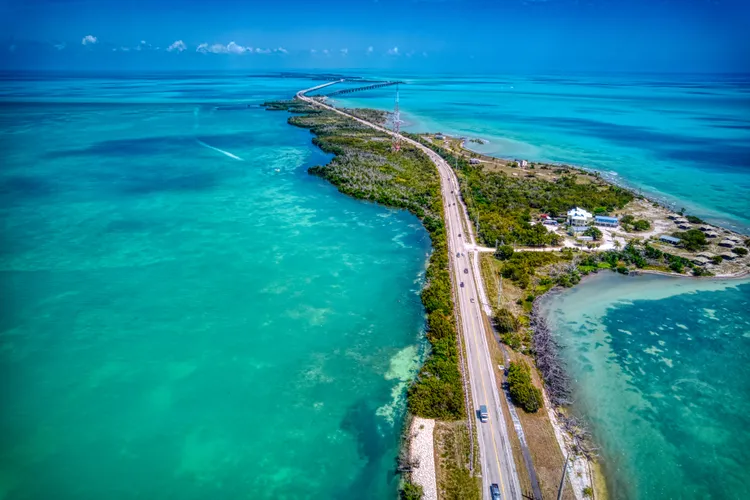 Aerial view of a long, narrow highway stretching over turquoise ocean waters, connecting small islands with lush greenery under a clear blue sky. Several cars travel along the scenic route.