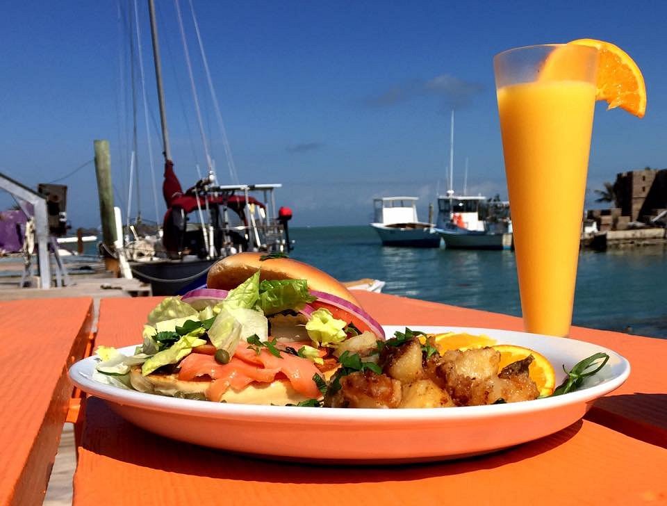 The ultimate marathon fl restaurants scene: from casual to fine dining 7 A plate with a burger, salad, and potatoes sits on an orange table by the water with boats docked in the background. A tall glass of orange juice with an orange slice garnish is beside the plate under a clear blue sky.