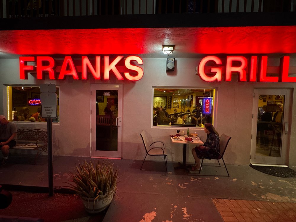 The ultimate marathon fl restaurants scene: from casual to fine dining 15 Nighttime view of franks grill with bright red neon letters. Two people dine at an outdoor table near large windows showing the restaurants interior. A glowing open sign is visible inside.