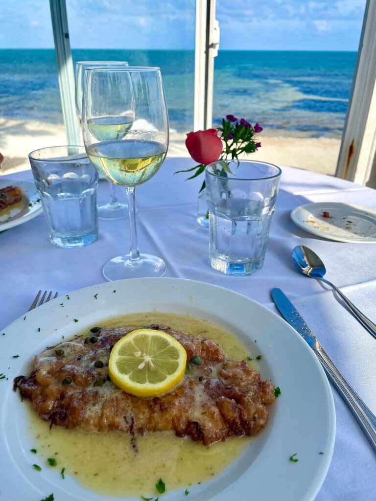 The ultimate marathon fl restaurants scene: from casual to fine dining 9 A plate of chicken piccata garnished with a lemon slice sits on a white tablecloth beside a glass of white wine, water glasses, and a small vase with flowers, overlooking the ocean through a window.