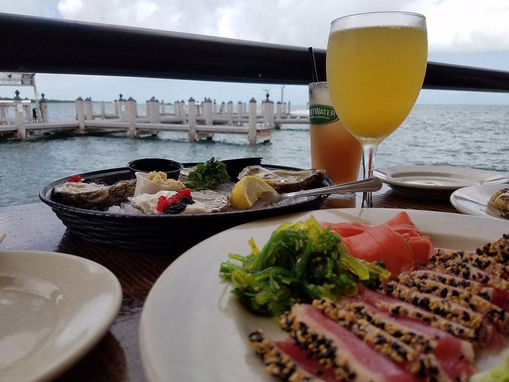 The ultimate marathon fl restaurants scene: from casual to fine dining 12 A table by the seaside holds plates of seared tuna with sesame, seaweed salad, oysters on ice with garnishes, and two drinks, with a dock and ocean visible in the background under a cloudy sky.