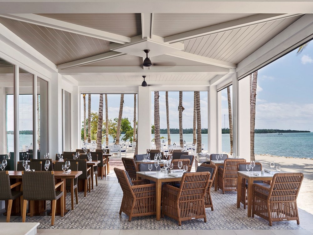 The ultimate marathon fl restaurants scene: from casual to fine dining 13 An open-air beachside restaurant with wicker chairs, wooden tables set with wine glasses, and a tiled floor. Palm trees and a sandy beach with calm blue water are visible outside.