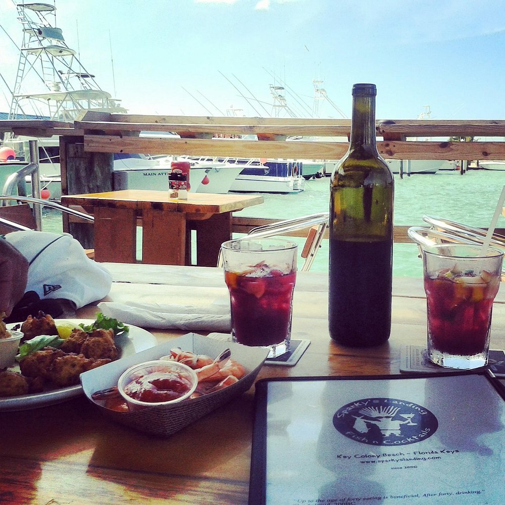 The ultimate marathon fl restaurants scene: from casual to fine dining 5 Two glasses of a dark drink and a wine bottle sit on a table next to a plate of seafood and sauce. In the background, fishing boats are docked by a turquoise sea under a sunny sky.
