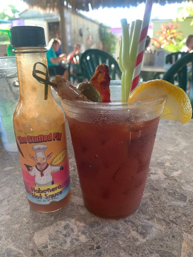 The ultimate marathon fl restaurants scene: from casual to fine dining 6 A plastic cup of bloody mary garnished with bacon, olives, lemon, and two striped straws sits on a table next to a bottle of the stuffed pig habanero hot sauce on an outdoor patio.
