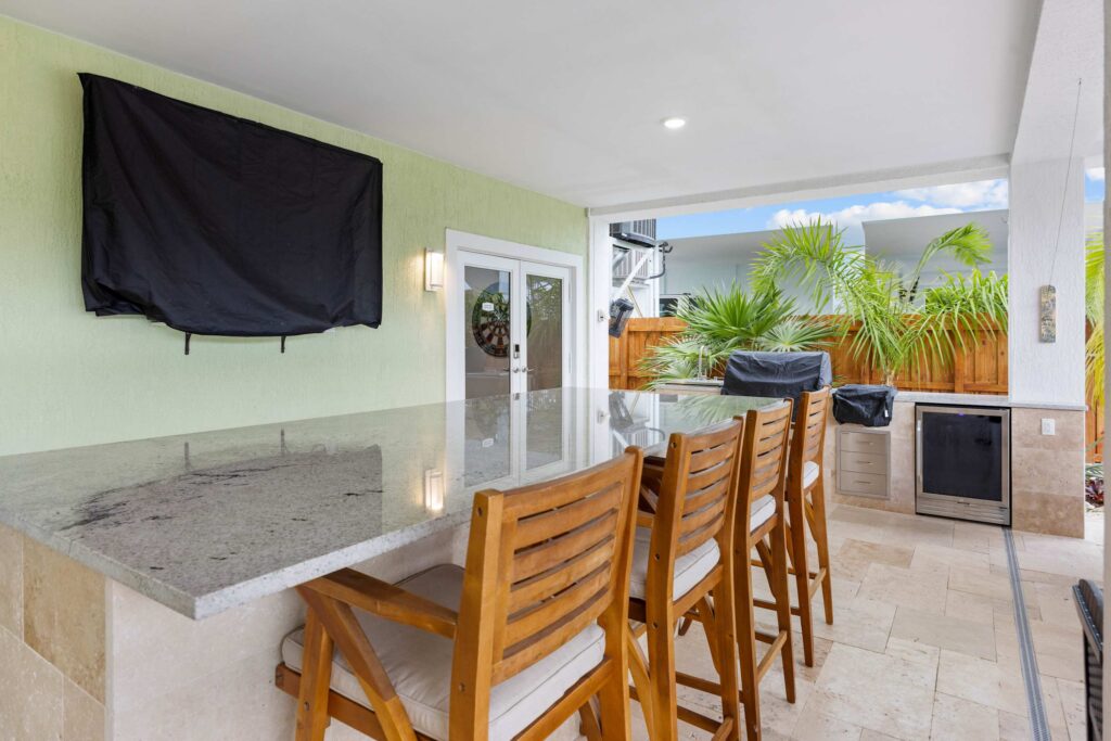 Outdoor kitchen with a granite countertop bar, four wooden chairs, a covered tv on the wall, a built-in grill, mini fridge, and tropical plants along a wooden fence in the background.