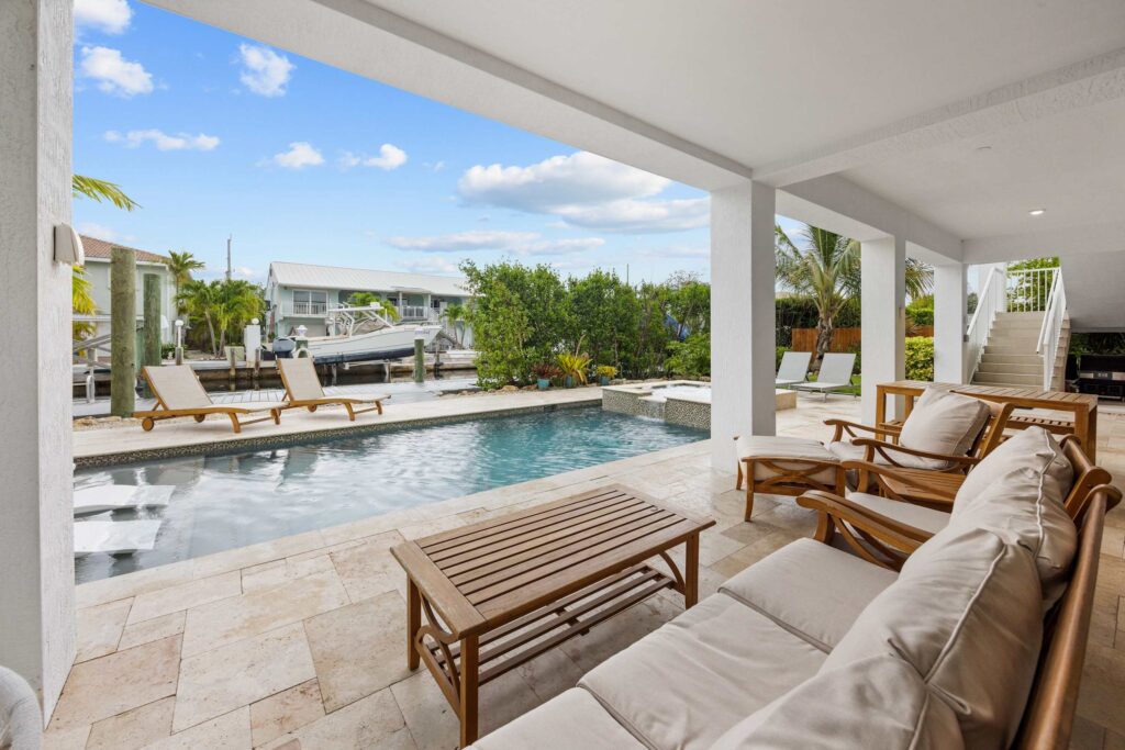 Covered patio with cushioned seating and wooden tables overlooks a pool with lounge chairs, set beside a canal with boats and surrounded by greenery and neighboring houses under a blue sky with clouds.