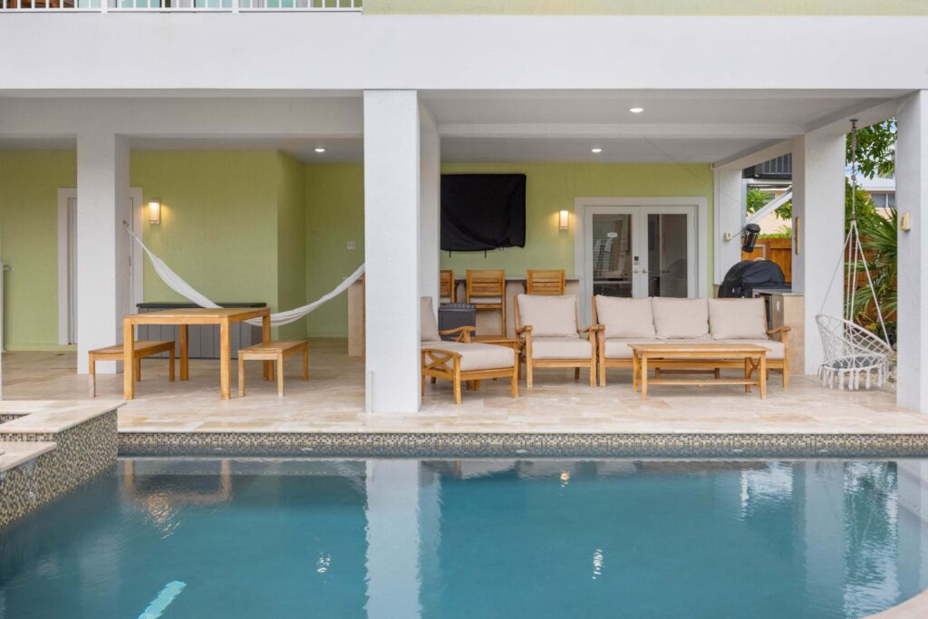 Covered patio area beside a swimming pool with cushioned wooden chairs, a table, a hammock, a wall-mounted tv, patio lights, and a hanging chair, all set against a light green wall.