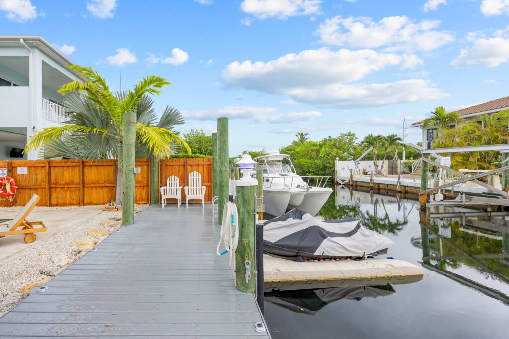 A waterfront dock with two white chairs, a palm tree, and a motorboat and jet ski tied nearby. The area is surrounded by fences, lush greenery, and residential buildings under a partly cloudy sky.