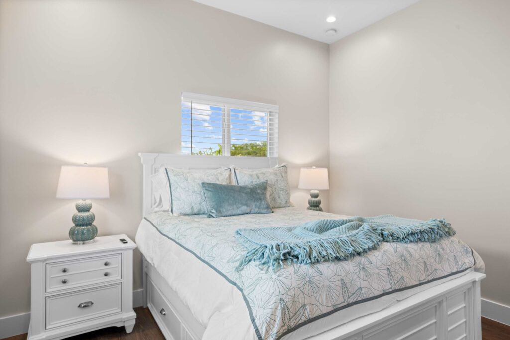 A bright bedroom with a white bed, light blue pillows and blanket, two matching bedside tables with lamps, light beige walls, and a window with white blinds letting in natural light.