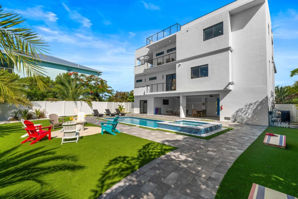 5-star vacation rental reviews: what actually earns them 3 A modern three-story white house with a spacious backyard featuring a pool, hot tub, lawn chairs in various colors, palm trees, and a paved walkway under a clear blue sky.