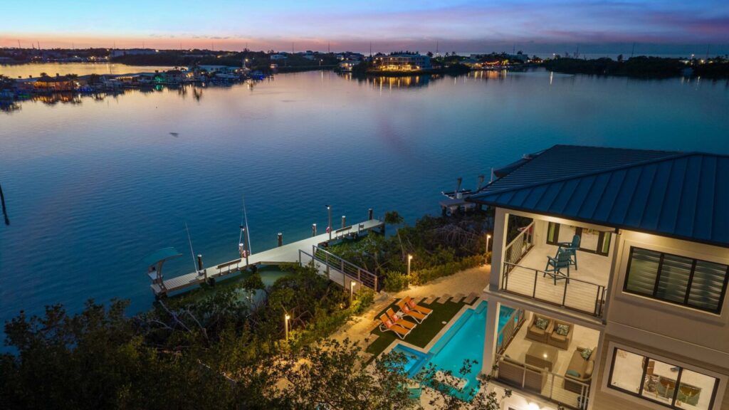 5-star vacation rental reviews: what actually earns them 6 A waterfront home with a lit pool and lounge chairs overlooks a calm bay at sunset, with boats docked nearby and houses and trees lining the water’s edge in the distance.