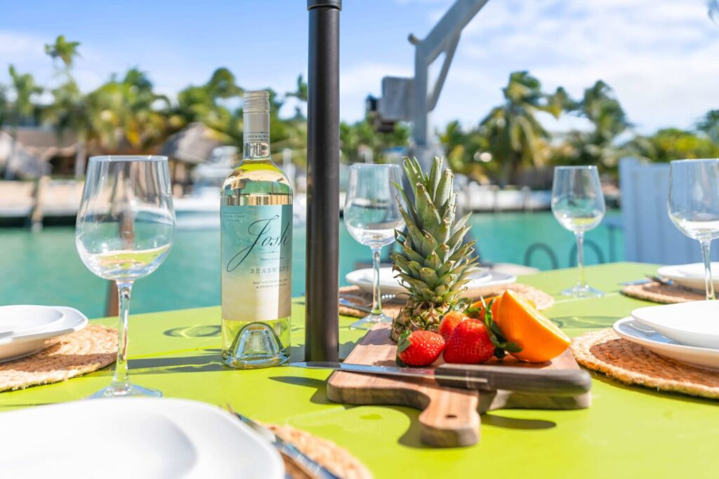 Florida keys happy hour: best overseas highway deals 6 A table set outdoors for a florida keys happy hour with wine, empty glasses, plates, and a cutting board holding pineapple, strawberries, and papaya overlooks sparkling water and swaying palm trees on a sunny day.