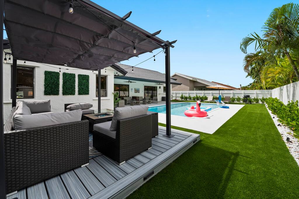 5 best restaurants in miami worth the trip | local picks 2026 8 Modern backyard with a shaded seating area, pool with inflatable floats, green artificial grass, and palm trees, adjacent to a white house on a sunny day.