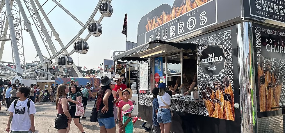 People gather at a churro food truck near a large ferris wheel at an outdoor fair. Some are ordering and others walk by, enjoying the sunny day and festive atmosphere.