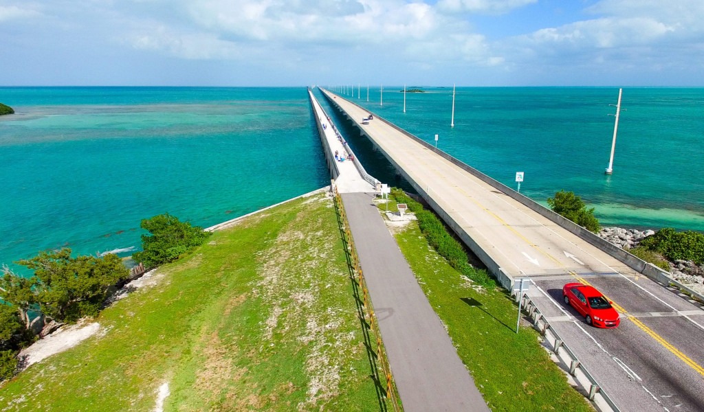 Aerial view of a long bridge stretching over turquoise ocean water, with a red car driving on the road. Lush greenery is visible at the start of the bridge, and the sky is partly cloudy.