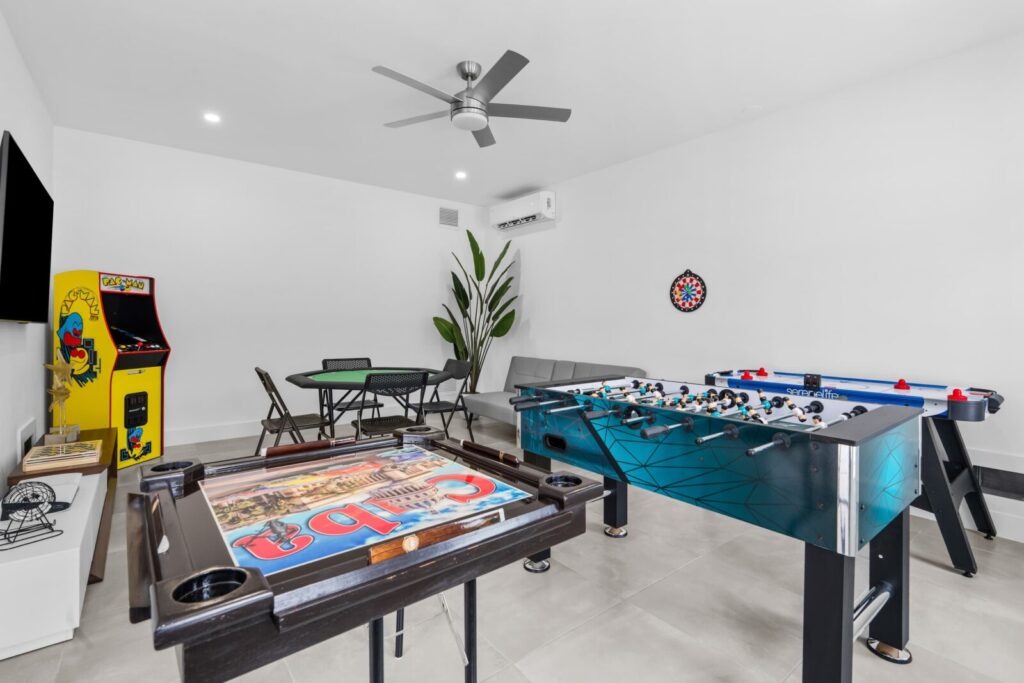 Modern game room with foosball table, pac-man arcade machine, poker table with chairs, dartboard on the wall, gray sofa, tv, and a large green plant in a bright, tiled space.