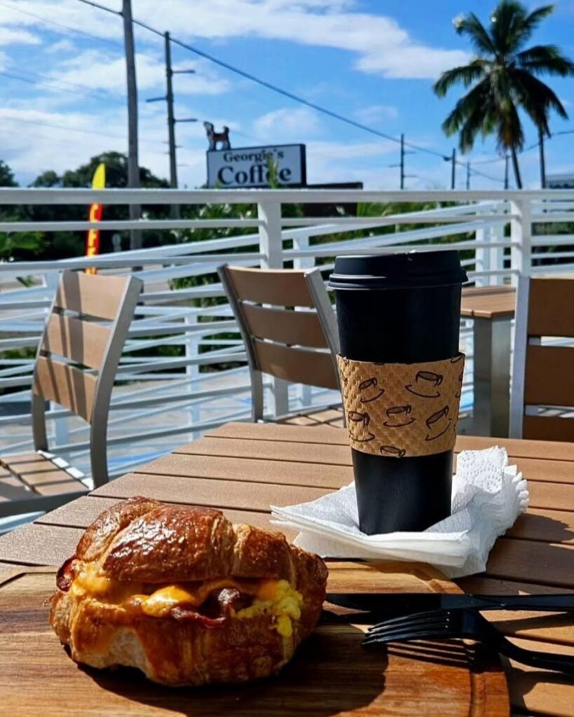 A croissant sandwich and a coffee cup sit on a wooden table outside a café. There are chairs, a railing, palm trees, a clear blue sky, and a coffee shop sign in the background.