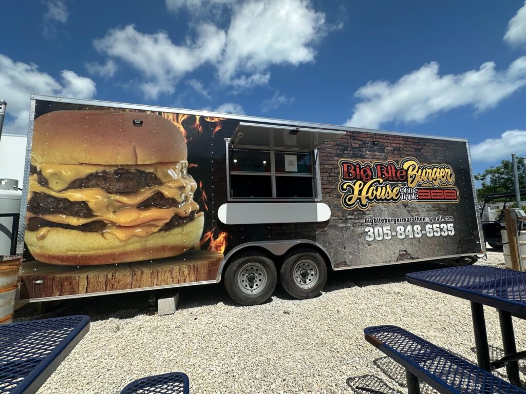 Food truck with a large image of a cheeseburger on the side, labeled big bite house burger barbecue, parked on gravel with a blue sky and picnic tables in the foreground. Phone number 305-848-6535 is displayed.
