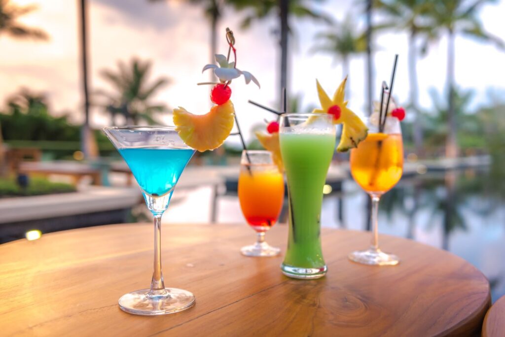 Florida keys happy hour: best overseas highway deals 2 Four colorful tropical cocktails with fruit garnishes sit on a wooden table beside a pool, evoking the vibrant spirit of a florida keys happy hour, with palm trees and a sunset in the blurred background.