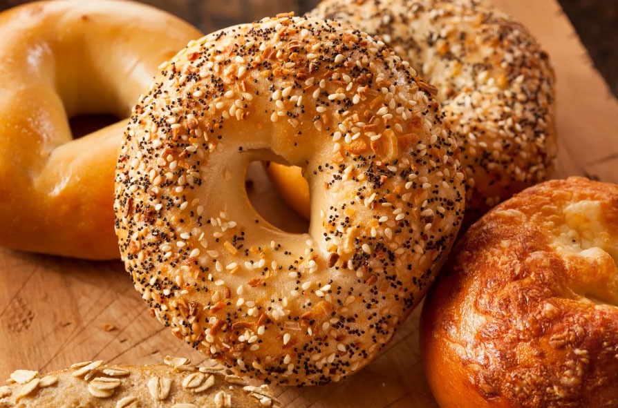 An assortment of bagels on a wooden surface, including everything, plain, oat-topped, and cheese varieties.