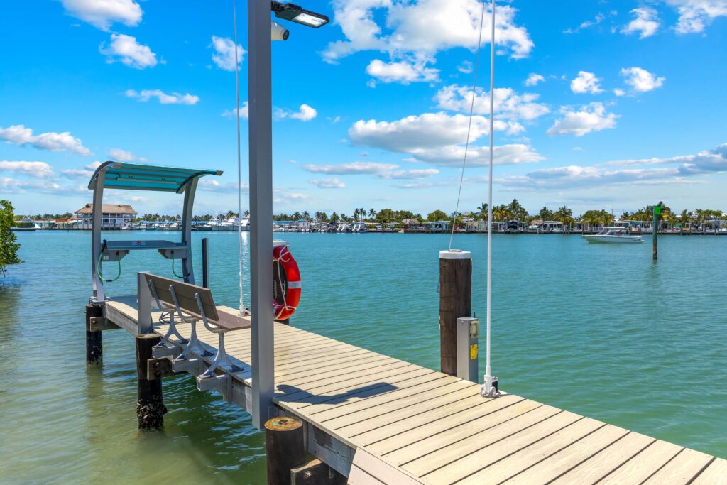 Do more vacation rental amenities mean more bookings? The truth (2026) 5 A wooden pier with benches and a shaded seat extends over calm blue water, with a red lifebuoy attached to a post. Boats and a marina are visible in the background under a bright, partly cloudy sky.