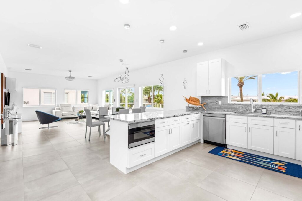 Bright, modern open-concept kitchen and living area with white cabinets, stainless steel appliances, marble countertops, large windows, and palm trees visible outside. Dining and lounge areas are in the background.