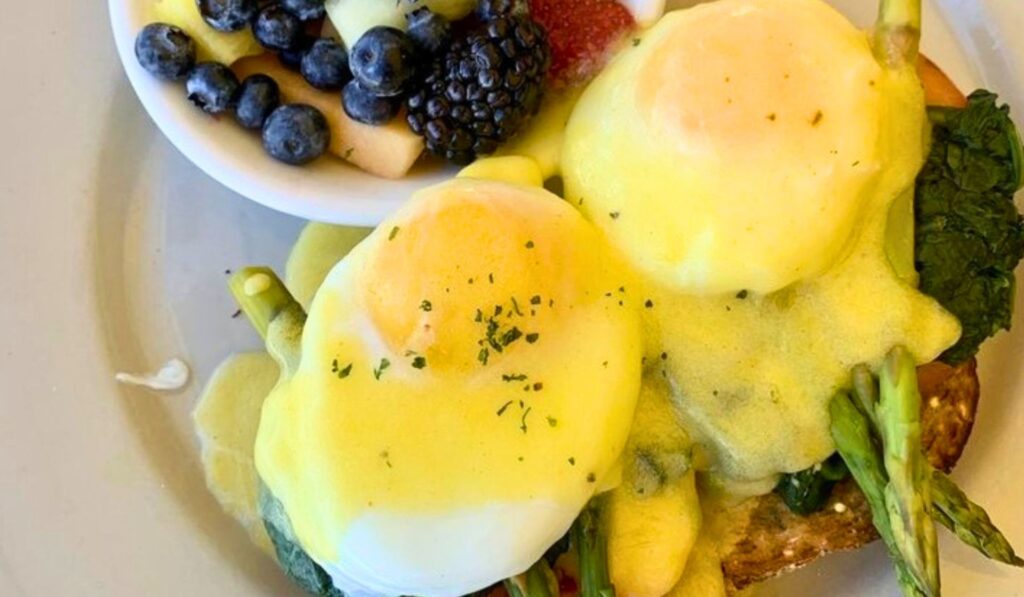 Two poached eggs topped with hollandaise sauce on toast with asparagus and spinach, served with a side bowl of mixed fruit including blueberries, blackberries, and apple slices.