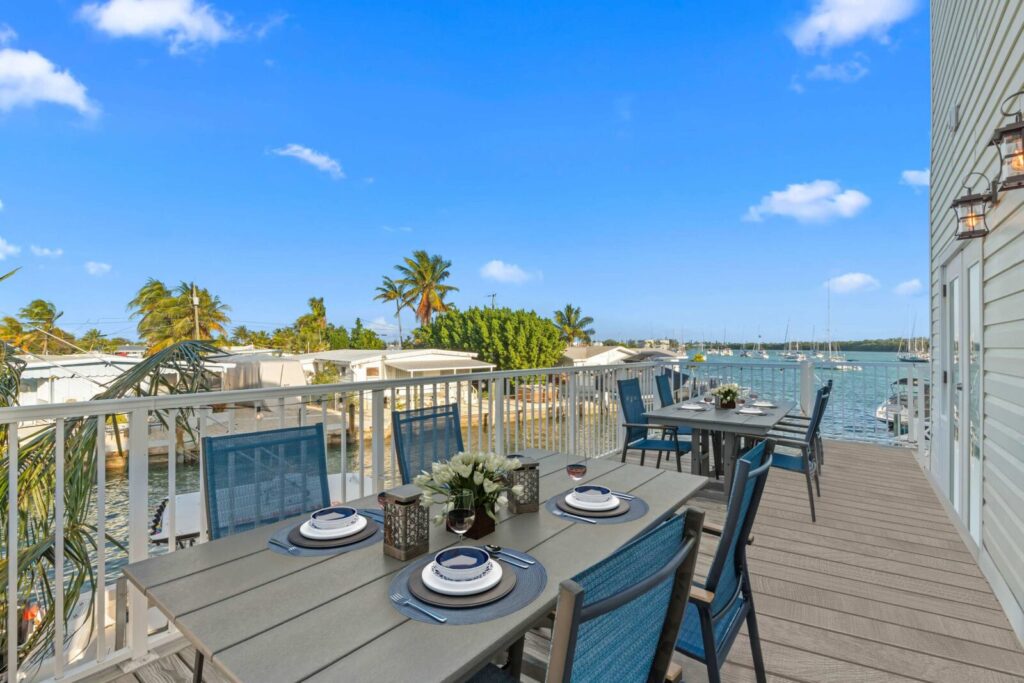 Outdoor dining area on a balcony overlooking a marina with boats. Tables are set with plates, glasses, and decorative flowers. Palm trees and blue sky are visible in the background.