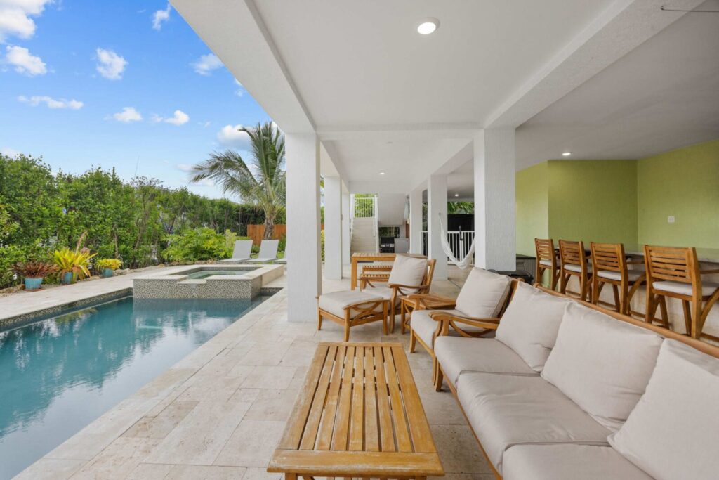 Modern outdoor patio with cushioned wooden furniture beside a rectangular swimming pool, surrounded by greenery and palm trees. A covered area includes a bar with wooden stools. Sky is clear with a few clouds.