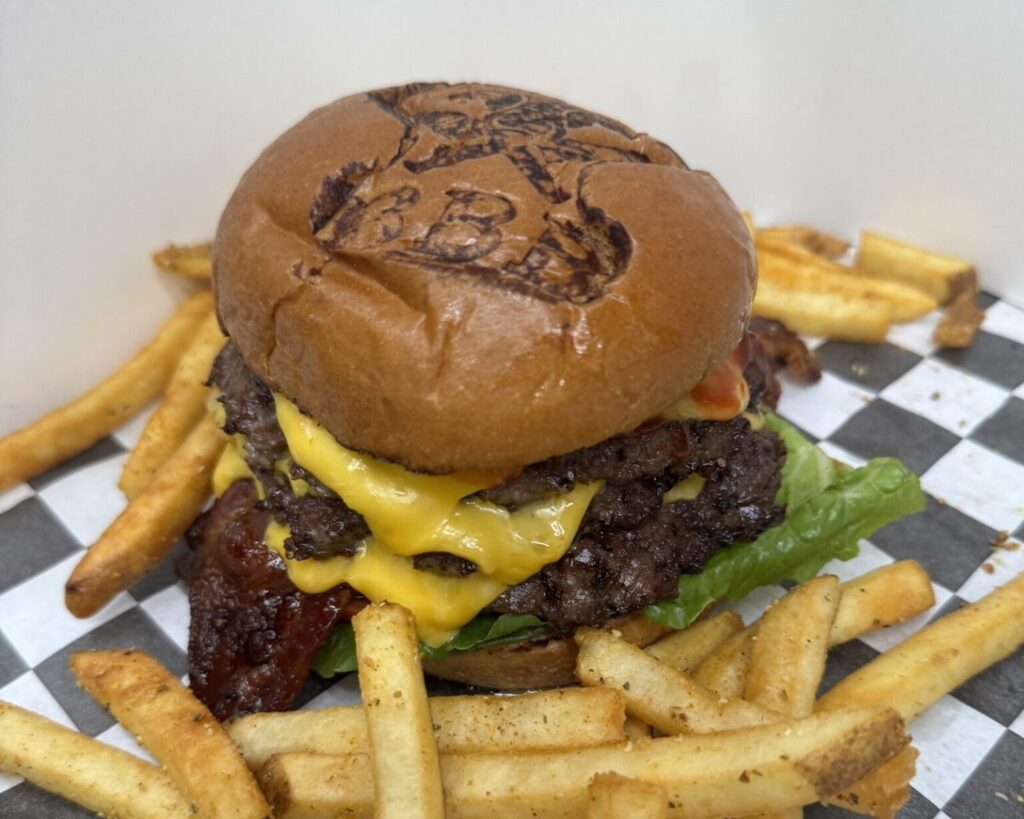 A double cheeseburger with lettuce, bacon, and two beef patties sits on checkerboard paper, surrounded by french fries. The top bun features a grilled design of a cartoon character.