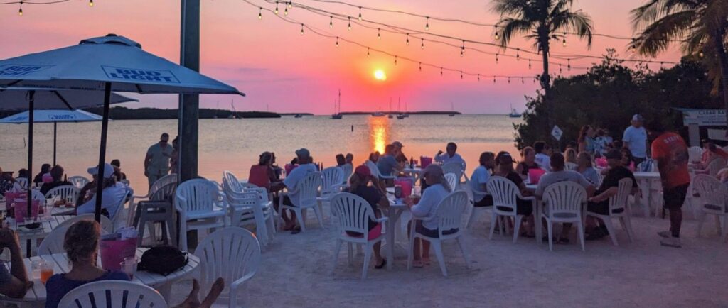 Florida keys happy hour: best overseas highway deals 3 People sit at white plastic tables and chairs on a sandy beach in the florida keys, watching the sunset over the water. String lights hang above, and palm trees and umbrellas add to the relaxed, festive atmosphere—perfect for enjoying happy hour by the shore.
