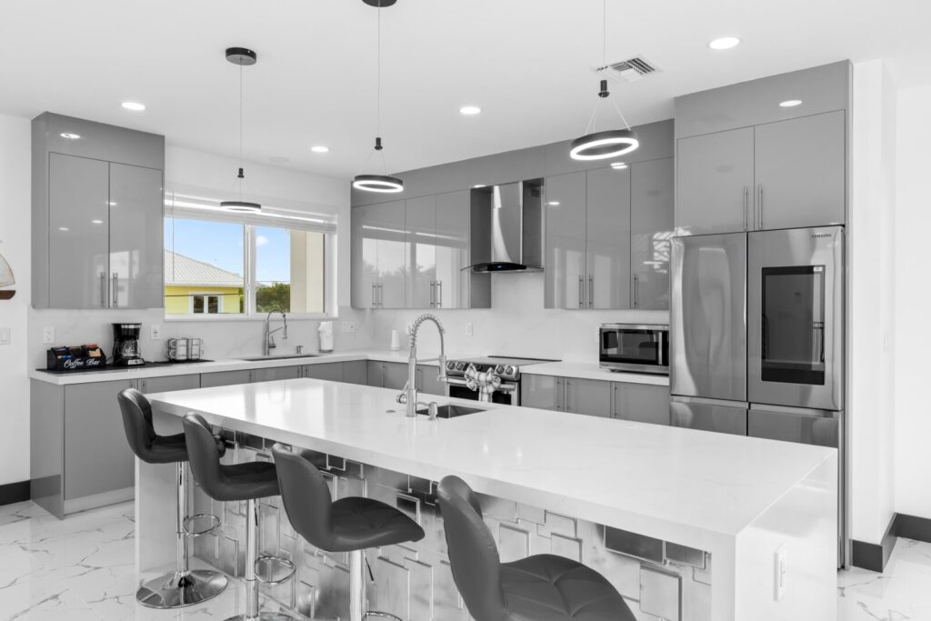 Do more vacation rental amenities mean more bookings? The truth (2026) 3 Modern kitchen with glossy gray cabinets, stainless steel appliances, built-in oven, and a large white island with sink and four black bar stools. Hanging pendant lights and a window bring in natural light.