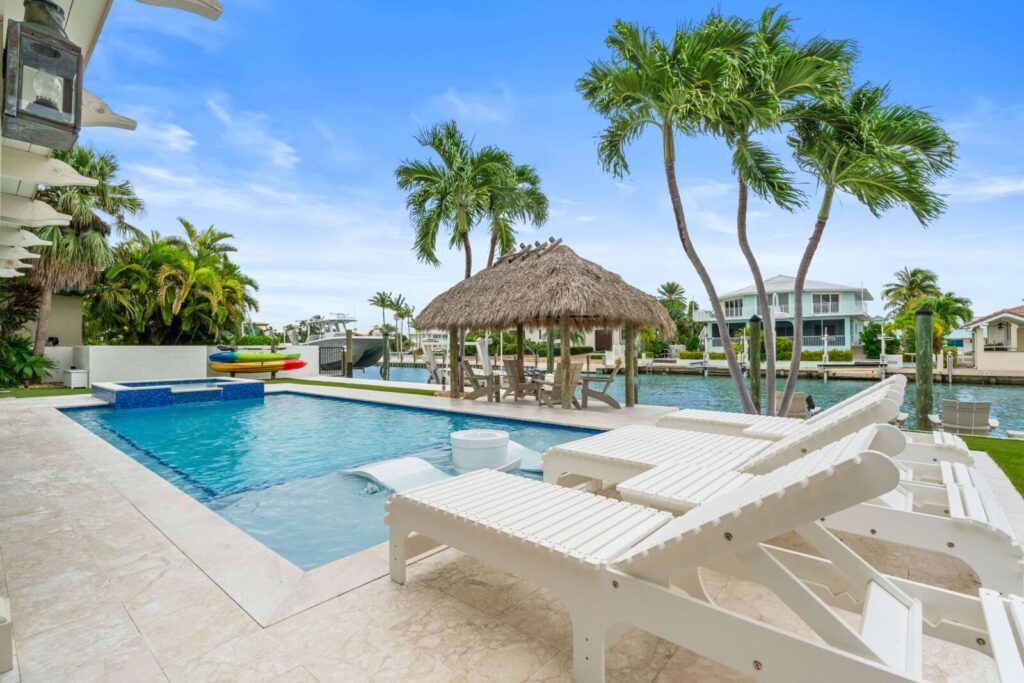 Do more vacation rental amenities mean more bookings? The truth (2026) 2 A tropical backyard with a swimming pool, white lounge chairs, a thatched-roof tiki hut, palm trees, and a canal with boats and waterfront houses in the background under a blue sky.
