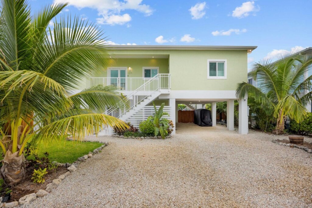 Light green raised house with white stairs, gravel driveway, tropical palm trees, and a covered parking area underneath the house. Blue sky with scattered clouds in the background.