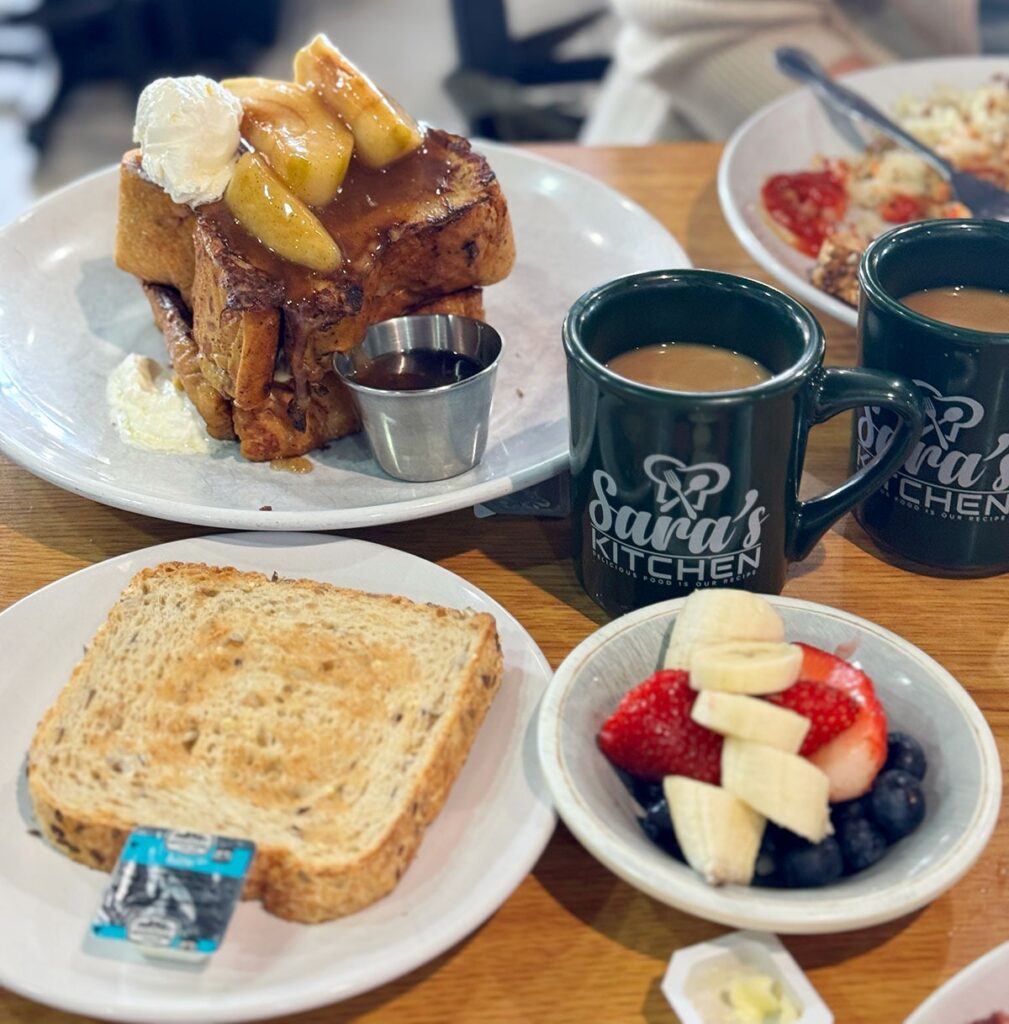 A breakfast spread with french toast topped with bananas, hot drinks in sara’s kitchen mugs, fruit salad, toast with butter, and dishes of eggs and tomato in the background.