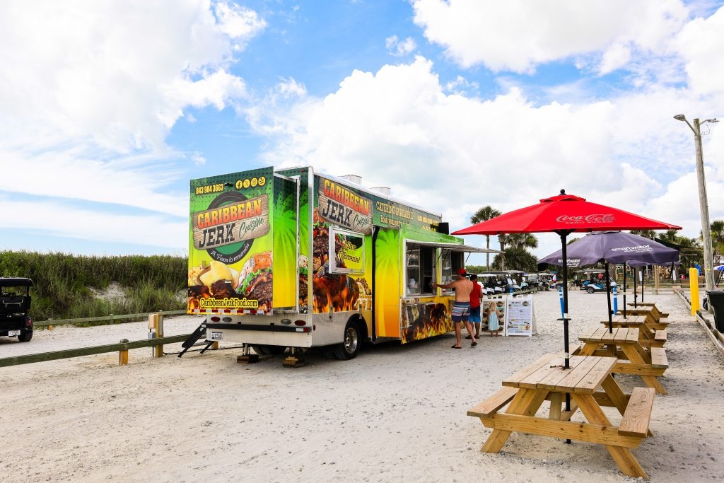 A colorful caribbean jerk food truck is parked on a sandy area near picnic tables with red and purple umbrellas. A customer stands at the window placing an order. Palm trees and a partly cloudy sky are in the background.