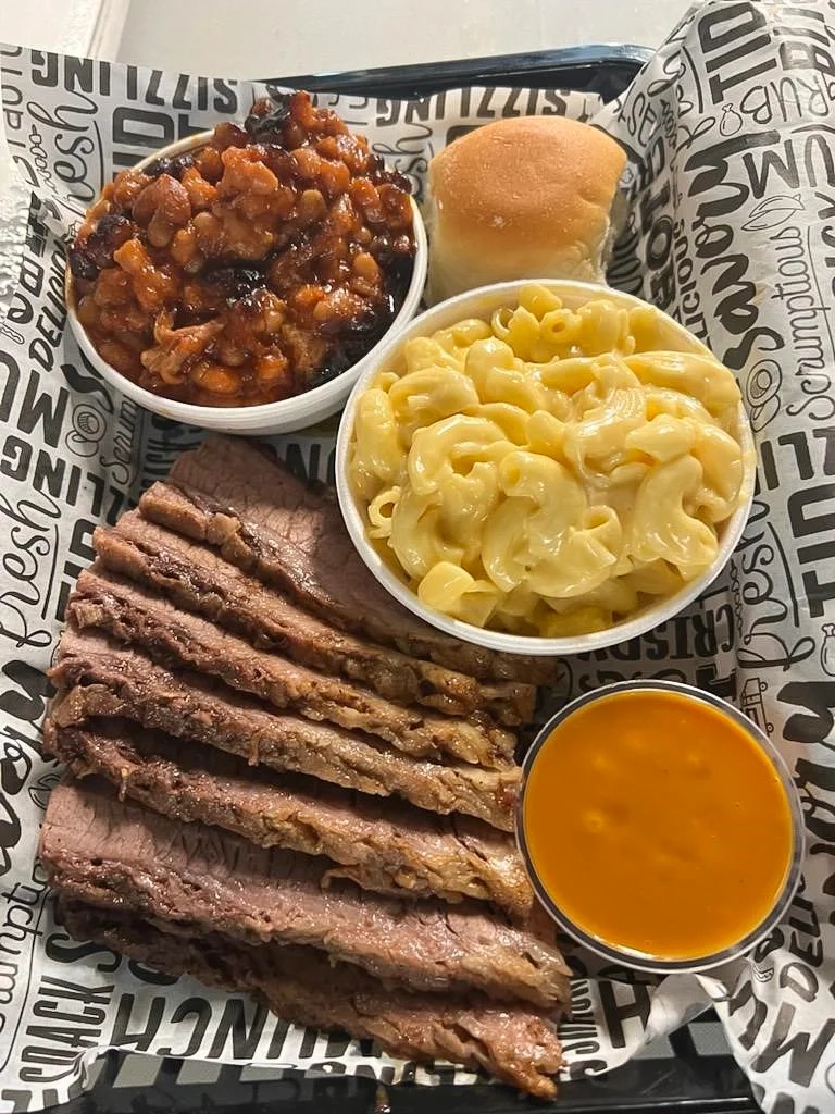 A tray with sliced brisket, macaroni and cheese, baked beans, a bread roll, and a cup of yellow barbecue sauce on patterned paper.