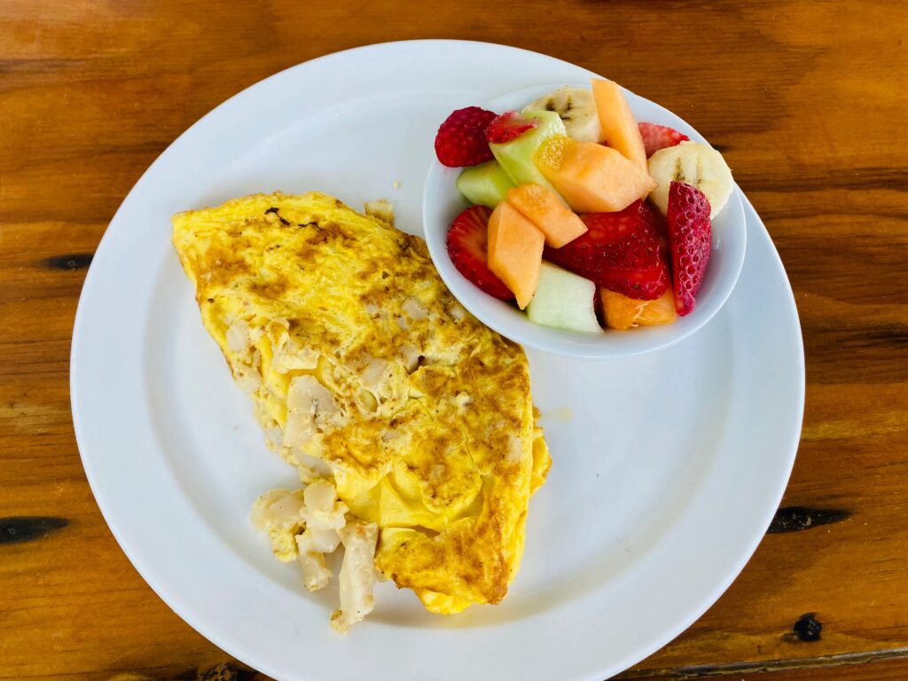 A plate with a plain omelette on one side and a small bowl of mixed fresh fruit, including strawberries, cantaloupe, banana, and honeydew, on a wooden table.