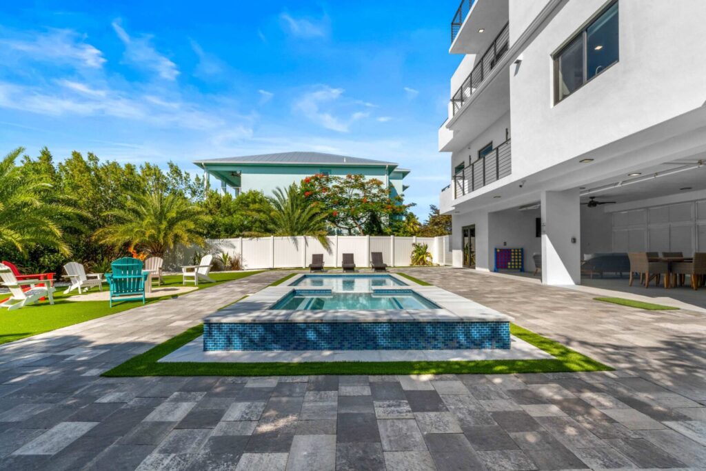 Do more vacation rental amenities mean more bookings? The truth (2026) 4 Modern backyard with a rectangular pool, hot tub, lounge chairs, and green artificial turf, surrounded by a white fence and palm trees, adjacent to a contemporary white house under a blue sky.